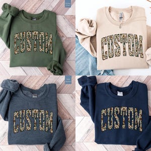 Custom Camo Shirt featuring bold varsity-style font — the perfect Camouflage Shirt for deer hunting, duck hunting, or casual wear during hunting season. This cozy Camo Sweatshirt makes a thoughtful Hunting Lover Gift, combining rugged outdoor style with personalized comfort.