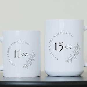 Puede incluir: Dos tazas de cer&aacute;mica blanca con texto negro y dise&ntilde;os florales. La taza m&aacute;s peque&ntilde;a dice "Custom Shirt and Gift Co 11 oz." y la taza m&aacute;s grande dice "Custom Shirt and Gift Co 15 oz."