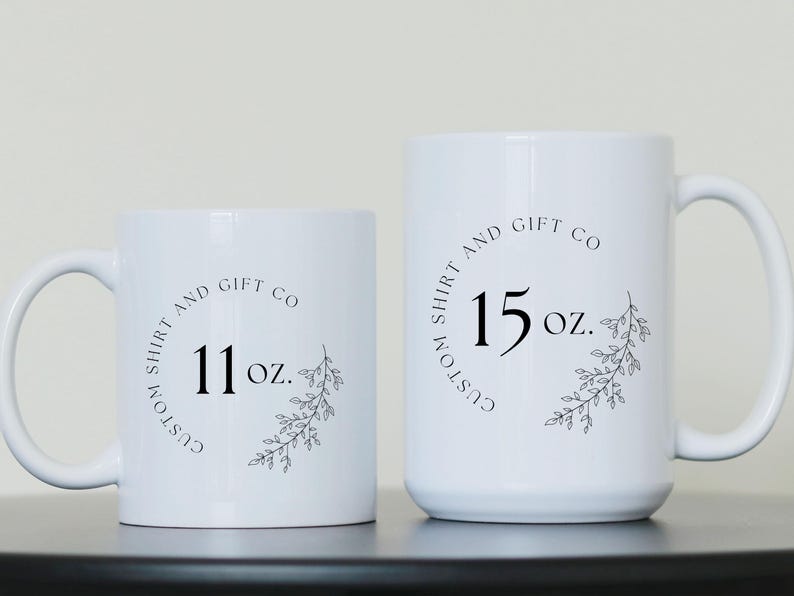 Puede incluir: Dos tazas de cer&aacute;mica blanca con texto negro y dise&ntilde;os florales. La taza m&aacute;s peque&ntilde;a dice "Custom Shirt and Gift Co 11 oz." y la taza m&aacute;s grande dice "Custom Shirt and Gift Co 15 oz."