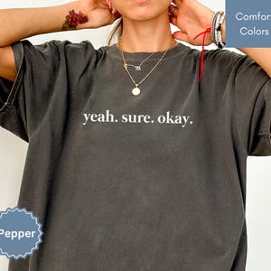 May include: A dark gray t-shirt with the text "yeah. sure. okay." printed in white on the front. The shirt is being worn by a person with a gold necklace.