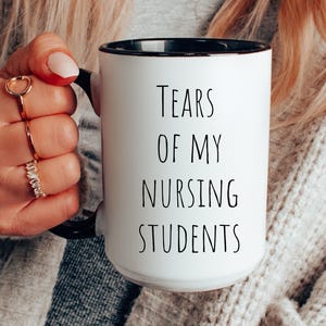 Könnte beinhalten: Eine weiße Keramik-Kaffeetasse mit schwarzem Rand und Henkel. Die Tasse hat einen schwarzen Schriftzug mit der Aufschrift "Tears of my Nursing Students".