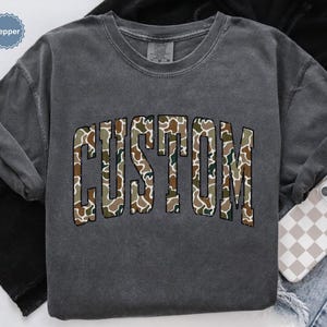 Custom Camo Shirt featuring bold varsity-style font — the perfect Camouflage Shirt for deer hunting, duck hunting, or casual wear during hunting season. This cozy Camo Sweatshirt makes a thoughtful Hunting Lover Gift, combining rugged outdoor style with personalized comfort.