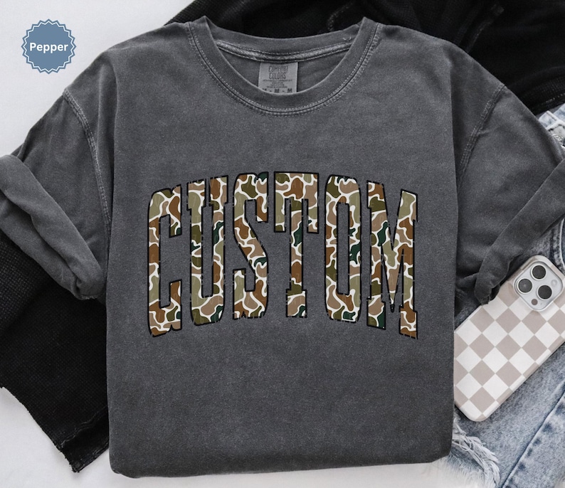 Custom Camo Shirt featuring bold varsity-style font — the perfect Camouflage Shirt for deer hunting, duck hunting, or casual wear during hunting season. This cozy Camo Sweatshirt makes a thoughtful Hunting Lover Gift, combining rugged outdoor style with personalized comfort.