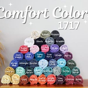 May include: A display of rolled Comfort Colors t-shirts in various colors, arranged in a pyramid shape. The image includes the text "Comfort Colors 1717" at the top. Colors include black, blue, green, pink, and yellow.