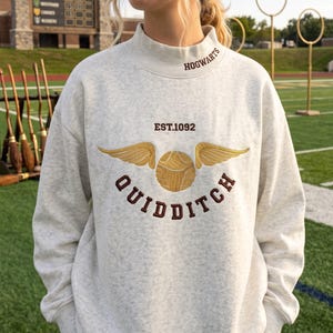 May include: A light gray heathered mock neck sweatshirt featuring embroidered text that reads HOGWARTS on the collar and EST.1092 QUIDDITCH below. The center design displays a golden snitch with detailed wings in gold thread and brown lettering.
