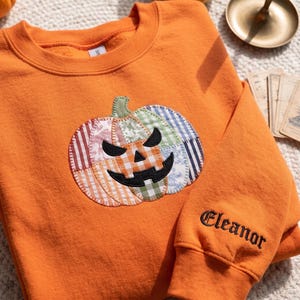 May include: An orange crewneck sweatshirt featuring a large, central pumpkin appliqué made from various fabric patches in plaid, striped, and floral patterns. The pumpkin has a black embroidered jack-o'-lantern face. The name Eleanor is embroidered in black on the sleeve.