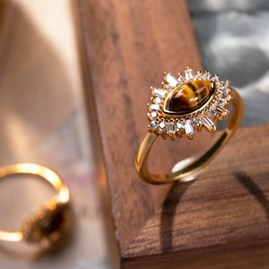 May include: A gold ring with a brown and gold gemstone surrounded by clear rhinestones. The ring is on a wooden surface.