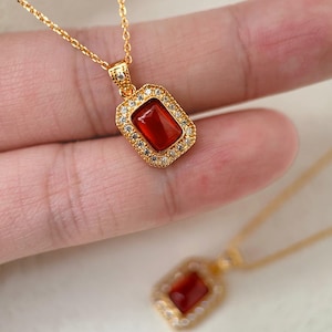 May include: Gold necklace with a red gemstone pendant surrounded by clear cubic zirconia stones. The pendant is in a rectangular shape with rounded corners.