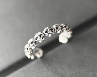 May include: Silver ring with a row of small skull designs. The ring is open and adjustable.