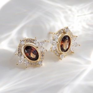 May include: A pair of gold earrings featuring a brown oval gemstone surrounded by smaller clear gemstones in a floral design.