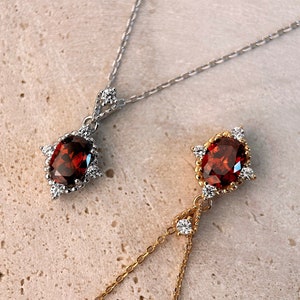 May include: Two necklaces with red gemstone pendants. The pendants are surrounded by smaller clear gemstones. One necklace has a silver chain and the other has a gold chain.