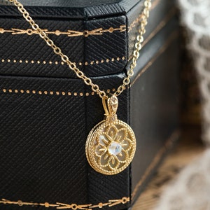 May include: A gold-colored necklace with a circular pendant featuring a floral design and a central gemstone. The pendant hangs from a delicate chain. The necklace rests on a black box with gold detailing, and a white lace fabric is in the background.