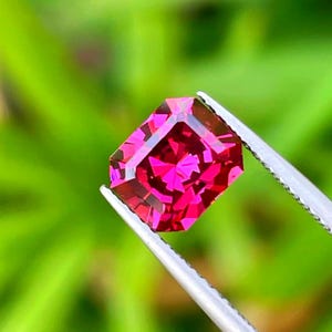 Puede incluir: Una gema de corte rectangular con un tono rojo magenta intenso y una superficie multifacetada, sostenida por unas pinzas de metal plateado. La piedra muestra bordes geométricos definidos y reflejos de luz internos en todo su cuerpo.