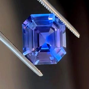 Op de afbeelding: Een vierkant geslepen edelsteen met facetten en een levendig blauw en violet kleurprofiel. De steen wordt vastgehouden door een zilverkleurige metalen pincet, wat de transparante helderheid en de geometrische slijpvorm met facetten benadrukt.