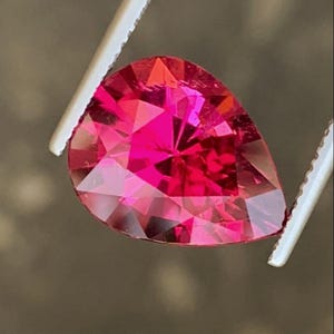 Puede incluir: Una piedra preciosa facetada en forma de pera con un color rojo rosado intenso y vibrante. La piedra presenta múltiples facetas triangulares y trapezoidales que reflejan la luz, creando una apariencia brillante y centelleante.