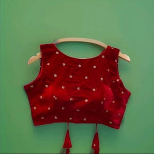 Puede incluir: Top sin mangas de terciopelo rojo con pequeñas y dispersas decoraciones doradas. El top tiene un escote redondo y está colgado de una percha de madera sobre un fondo verde. Borlas rojas cuelgan del dobladillo inferior.