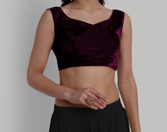 May include: A sleeveless, plum-colored velvet crop top with a scoop neckline. The top is paired with a black skirt. A gold-toned ring with a square design is worn on the hand.