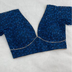 Puede incluir: Top azul oscuro con cuello en V, estampado de hojas y detalles de lentejuelas. Los bordes están ribeteados con un borde festoneado dorado claro. El top está hecho de una tela suave y fluida.