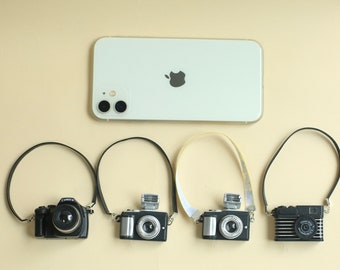 May include: Four miniature cameras with black straps hanging against a white background. The cameras are all different styles and have black and silver accents.