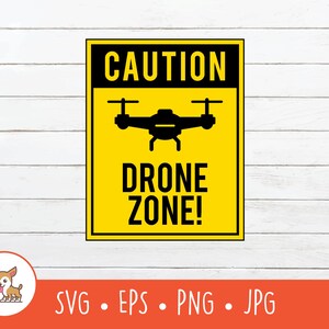 May include: A yellow and black sign with the words "CAUTION" and "DRONE ZONE!" in bold, black text. A black silhouette of a drone is in the center. The sign is on a white wooden background.