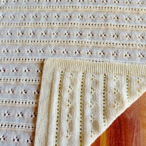 May include: Cream-colored knitted blanket with a detailed openwork pattern. The blanket features horizontal rows of small, decorative holes and vertical columns of textured stitches. The corner is folded over, revealing the intricate knit design. The blanket is made of soft yarn.