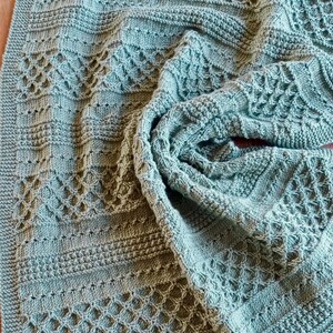 May include: A knitted blanket in a soft, muted teal color. The blanket features a variety of textured patterns, including diamond and horizontal stripe designs. The edges are neatly finished with a ribbed border, creating a cozy and handcrafted feel.