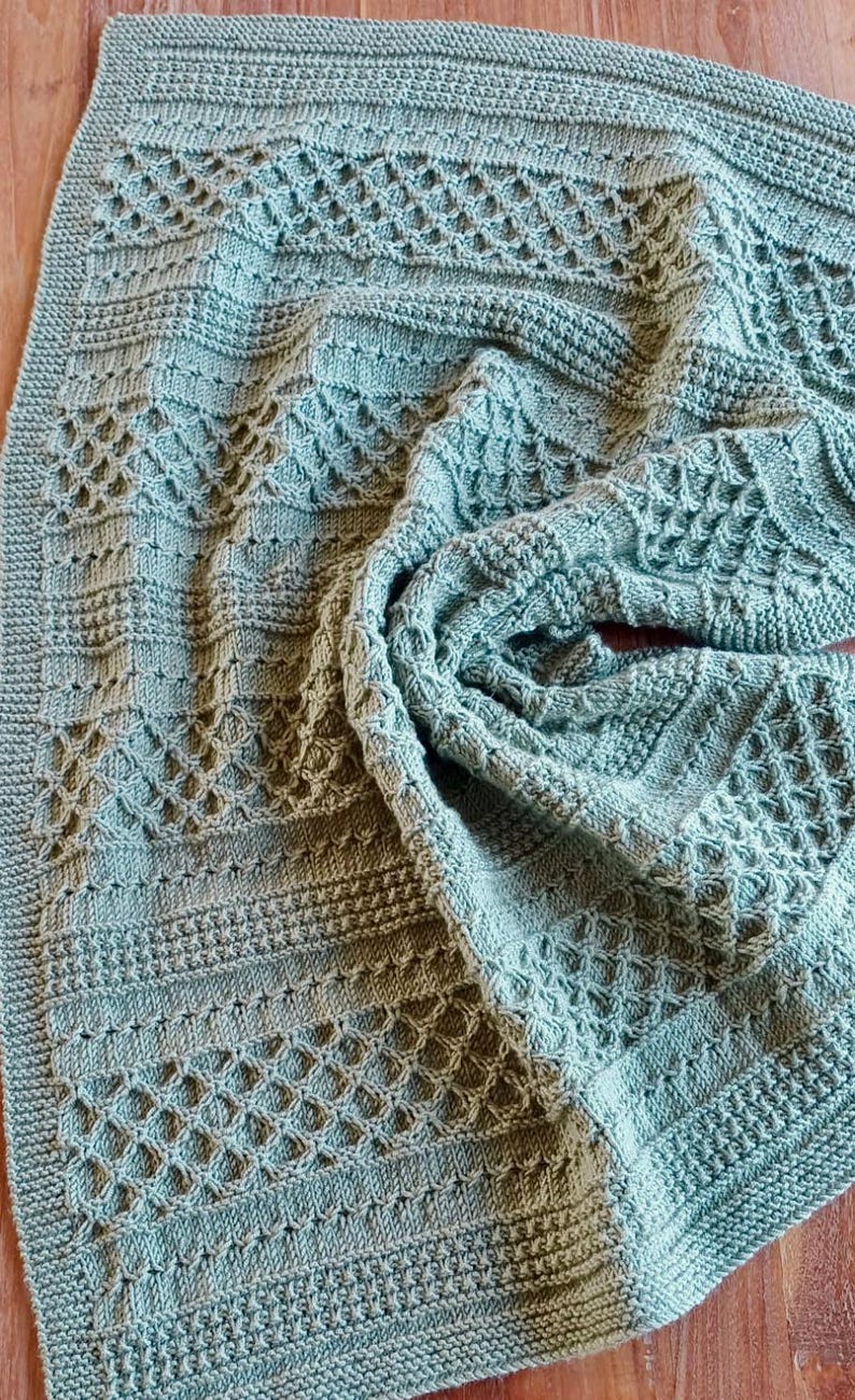 May include: A knitted blanket in a soft, muted teal color. The blanket features a variety of textured patterns, including diamond and horizontal stripe designs. The edges are neatly finished with a ribbed border, creating a cozy and handcrafted feel.