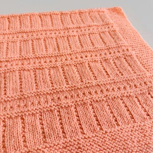 May include: A square-shaped, hand-knitted blanket in a solid peach color. The textile features a repeating pattern of vertical ribbed columns separated by horizontal rows of garter stitch, with a wide garter stitch border framing the edges.