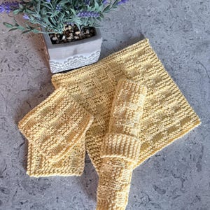 May include: A set of three pale yellow knitted washcloths featuring a textured basketweave pattern. The set includes one large square cloth, one smaller square cloth, and one rectangular cloth rolled into a cylinder shape.