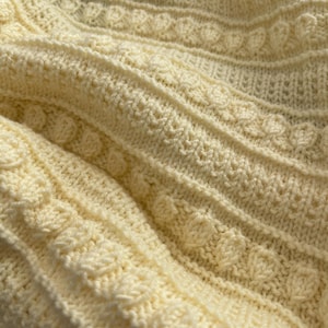 Puede incluir: Primer plano de una manta de punto de color crema con un patrón texturizado. La manta está hecha de hilo suave y tiene un diseño delicado e intrincado.