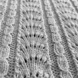 May include: A white knitted fabric featuring a repeating pattern of vertical columns. The design alternates between wide panels of openwork fan-shaped lace stitches and narrow, raised vertical bands of textured, rounded cable-like knit stitches.