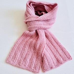 Puede incluir: Una bufanda de punto de color rosa claro con una textura acanalada vertical constante a lo largo de toda la tela. La bufanda está doblada sobre sí misma, mostrando un patrón de puntadas denso y uniforme con bordes limpios.