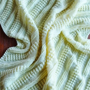 Peut inclure: Une couverture tricotée de couleur crème avec un motif texturé. La couverture présente des motifs diagonaux côtelés et en forme de losange. Le tissu est plié, créant des ombres et mettant en valeur le travail de tricot. La couverture est en matière douce et chaude.
