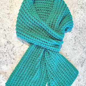 May include: A teal-colored, hand-crocheted scarf. The scarf is made of a soft yarn and features a textured stitch pattern. The scarf is folded and draped, showcasing its length and design. The scarf is a versatile accessory for any wardrobe.