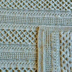May include: Close-up of a knitted blanket in shades of green. The blanket features various textured patterns, including diamond and ribbed designs. The corner of the blanket is folded over, revealing the detailed edge. This handmade item is perfect for home decor.