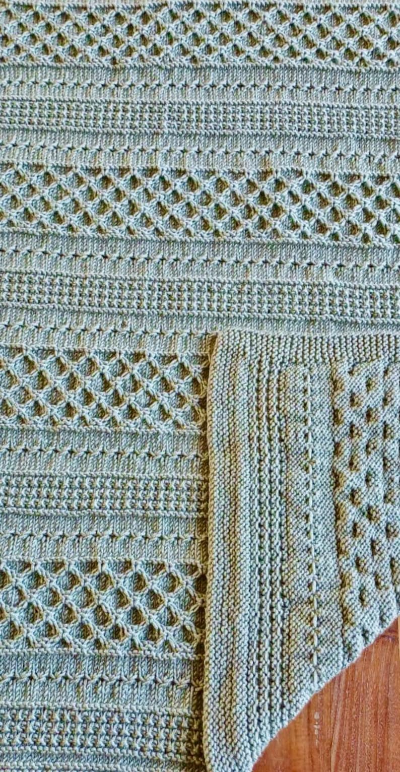 May include: Close-up of a knitted blanket in shades of green. The blanket features various textured patterns, including diamond and ribbed designs. The corner of the blanket is folded over, revealing the detailed edge. This handmade item is perfect for home decor.