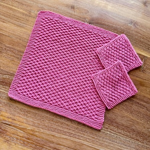 May include: A set of three square, hand-knitted washcloths in a vibrant magenta pink color. Each piece features a consistent, textured diamond-like stitch pattern throughout, with a simple garter stitch border framing the edges of the fabric.