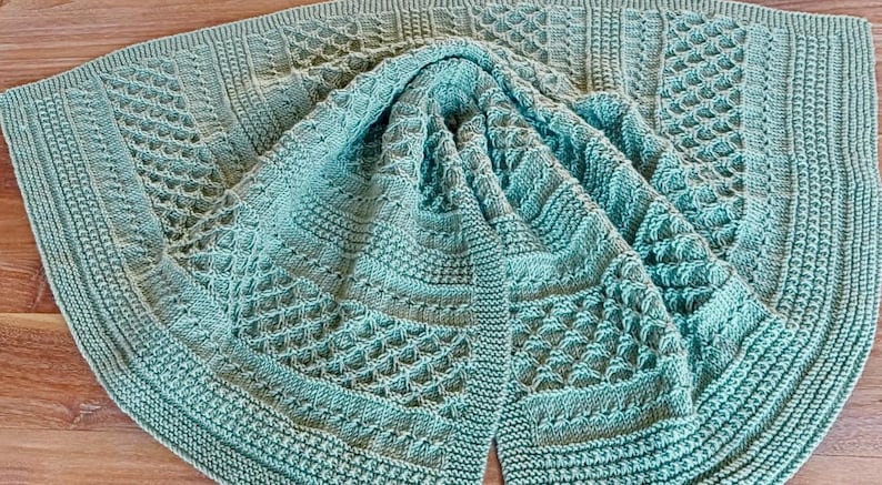 May include: A folded, teal-colored knitted blanket with intricate patterns. The blanket features various textured designs, including diamond and geometric shapes, and a ribbed border. The soft, folded fabric is displayed on a wooden surface.