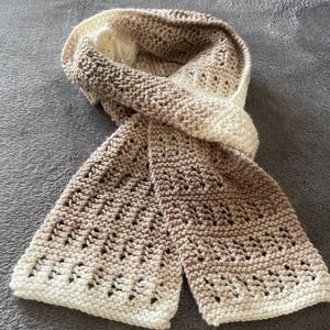 May include: A knitted scarf with a gradient of beige and white colours. The scarf has a lacy pattern and is folded in half.