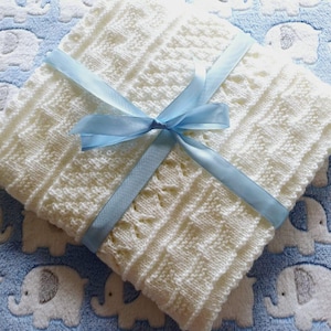 Può includere: Una coperta lavorata a maglia bianca piegata con un motivo strutturato di strisce verticali e forme a rombo. La coperta è legata con un nastro di raso azzurro liscio, annodato in un fiocco al centro del tessuto piegato.