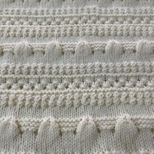 May include: Cream-colored knitted blanket with a textured pattern. The blanket features rows of raised, rounded shapes and various stitch patterns, creating a visually interesting and tactile surface. The overall design is simple and elegant.