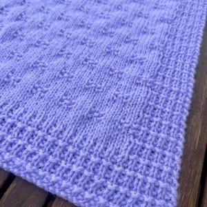 May include: A close-up of a lavender knitted blanket. The blanket features a textured pattern with a ribbed border. The soft, pastel color and detailed stitching create a cozy and handcrafted appearance. The blanket is made of a soft yarn.