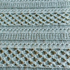 May include: Close-up of a knitted textile in shades of blue and brown. The pattern features a combination of horizontal stripes and diamond-shaped openwork designs, creating a textured surface. The yarn appears soft and the overall aesthetic is cozy.