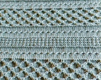 May include: Close-up of a knitted textile in shades of blue and brown. The pattern features a combination of horizontal stripes and diamond-shaped openwork designs, creating a textured surface. The yarn appears soft and the overall aesthetic is cosy.