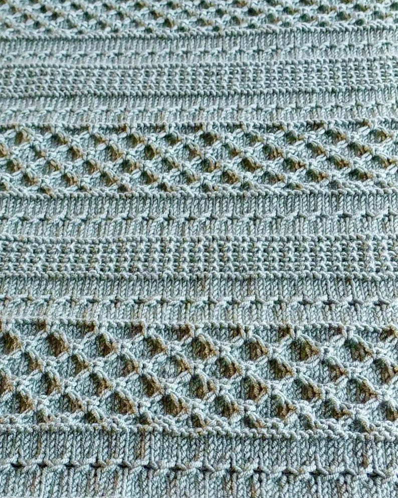 May include: Close-up of a knitted textile in shades of blue and brown. The pattern features a combination of horizontal stripes and diamond-shaped openwork designs, creating a textured surface. The yarn appears soft and the overall aesthetic is cozy.