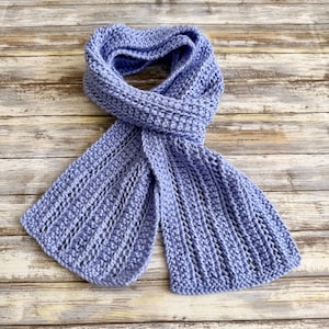 May include: A light purple crochet scarf featuring a textured ribbed pattern with vertical stitches. The scarf is constructed with a soft yarn material and displays a consistent knit design throughout the entire length of the accessory.