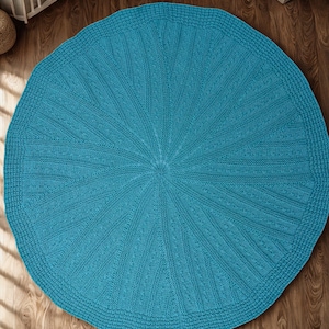 May include: A round, turquoise knitted blanket with a textured pattern. The blanket has a central design that radiates outwards, with a detailed border. The blanket is approximately 60 inches in diameter.