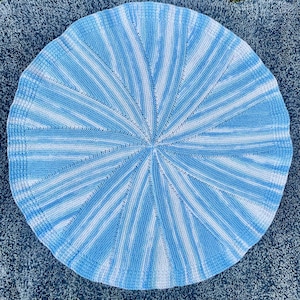 May include: A circular knitted blanket in light blue and white. The design has radiating stripes from the centre, forming a starburst pattern. The blanket is made of soft yarn and has a textured appearance.