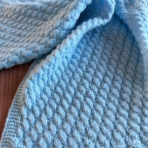 May include: A light blue knitted blanket featuring a repeating diagonal cable stitch pattern. The textile has a textured surface with a consistent weave throughout, finished with a simple garter stitch border along the bottom edge of the fabric.