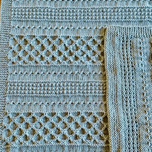 May include: A close-up of a light blue knitted blanket with various textured patterns. The blanket features rows of horizontal ribbing, diamond-shaped openwork, and a textured border. The corner of a smaller, matching square is visible.